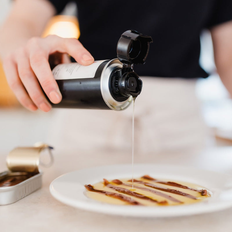 A person in a black shirt pours olive oil from a black and silver bottle onto a white plate with anchovies, in a brightly lit kitchen setting.