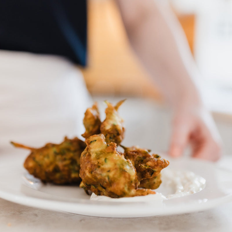 Golden-brown fried fritters served on a white plate with yoghurt, with a blurred person's hand and torso in the background.