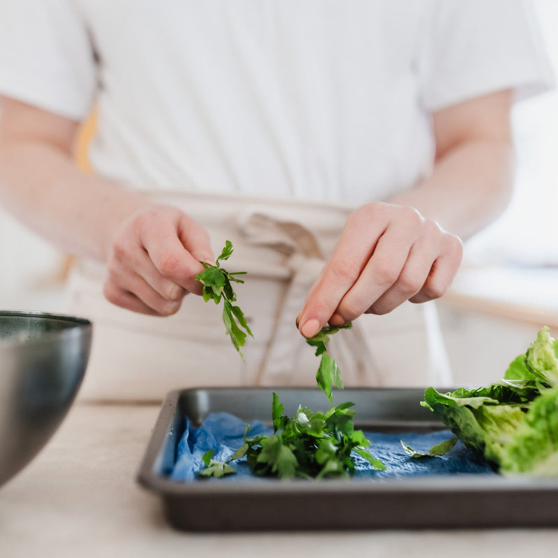 A person in a white shirt and apron preparing fresh green herbs over lettuce on a baking sheet, with a metal bowl in the foreground.