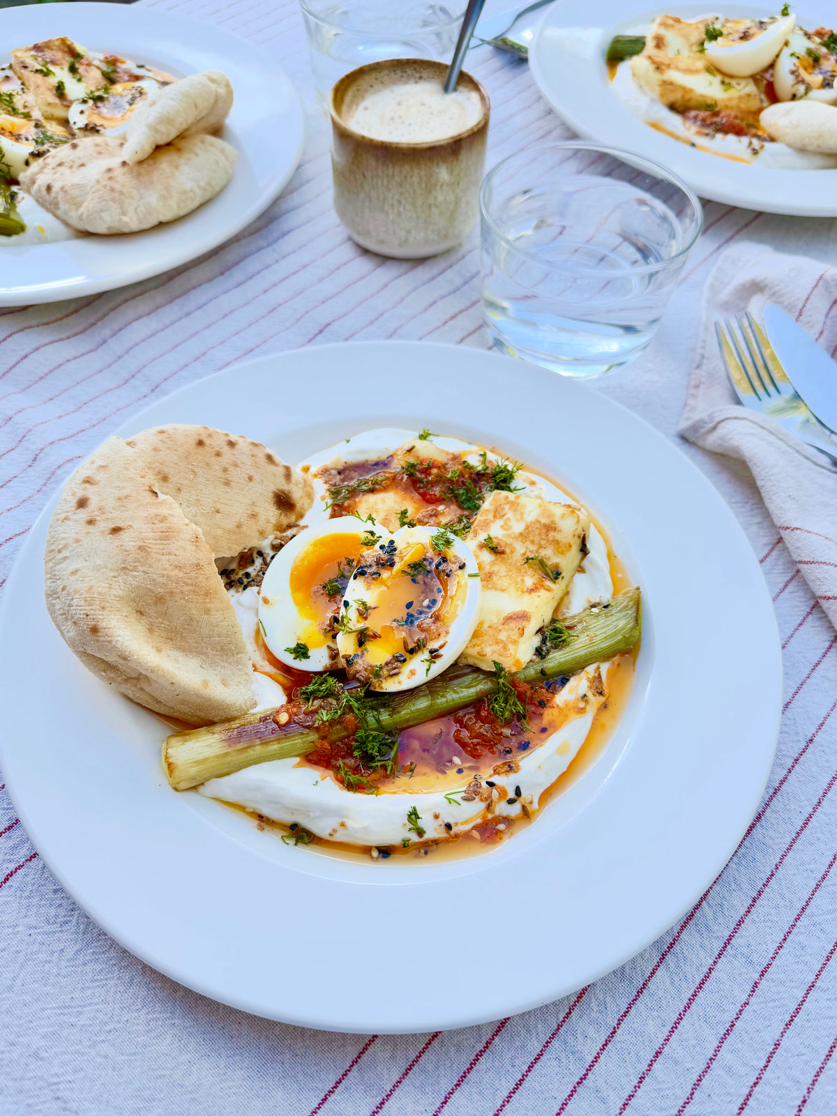 A plated breakfast dish on a white plate, featuring a poached egg with a runny yolk, baby leek, and flatbread on a striped tablecloth, with other plates and a drink blurred in the background.