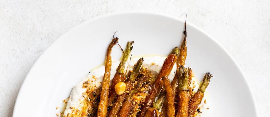 Roasted carrots on yoghurt with a drizzle of oil and sprinkle of dukkah, served on a white plate against a light textured background.
