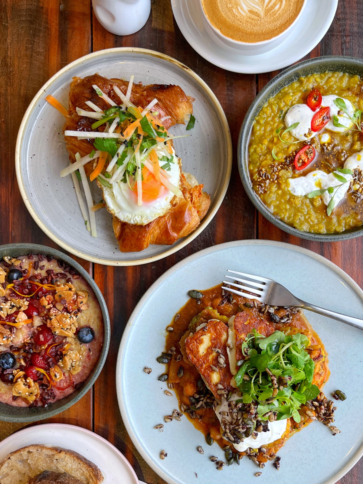 Assorted dishes including a fried egg on a kimchi and cheddar croissant, a bowl of dahl, porridge and a plate with halloumi, egg and fritters on a wooden table.